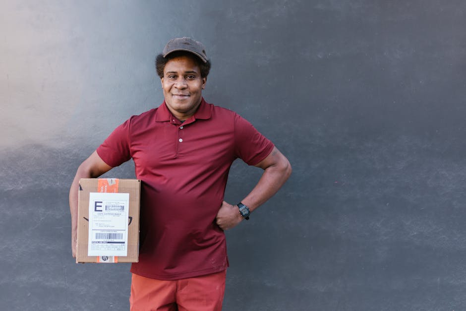 A smiling delivery person with medium brown skin, wearing a dark gray cap, a maroon polo shirt, and matching red pants, stands outdoors against a plain gray wall. He is holding a small, rectangular ca