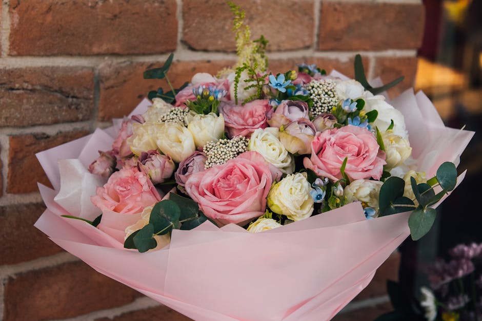 A floral bouquet arranged with pink roses, white lisianthus, small blue forget-me-nots, and sprigs of white baby's breath, surrounded by green eucalyptus leaves. The flowers are wrapped in soft pink t