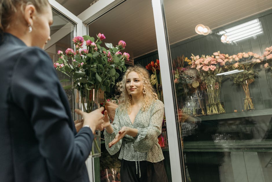 A woman with curly blonde hair and light skin, dressed in a light-colored blouse, is receiving a bouquet of fresh pink roses from a florist inside a flower shop. The florist, whose face is not visible