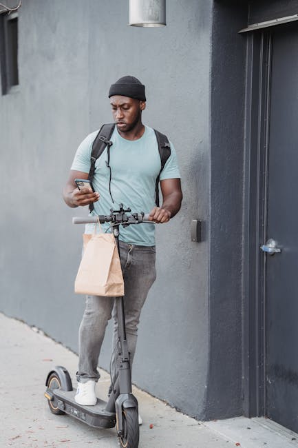 A young man with dark skin, wearing a black beanie, light grey T-shirt, and grey jeans, is standing outdoors against a grey wall. He is holding a smartphone in his right hand and a brown paper shoppin